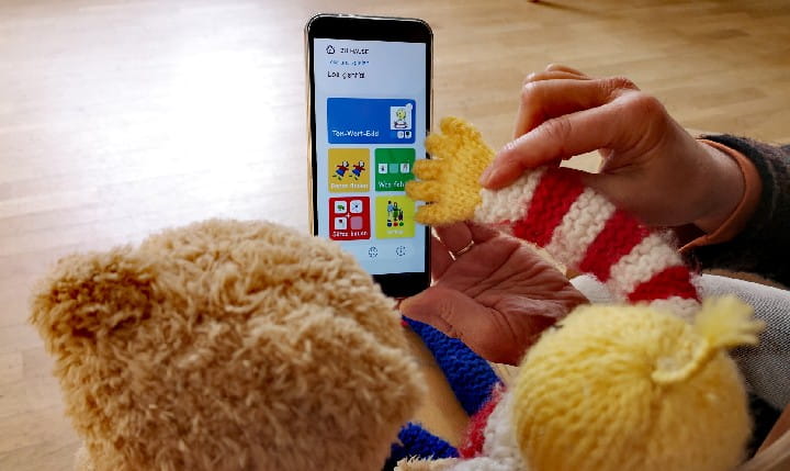 Eine Puppe und ein Teddybär sitzen vor einem Handy mit der Kikus App