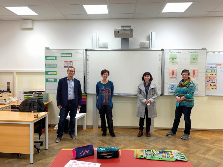 Vier Personen stehen vor einer interaktiven Tafel in einem Klassenzimmer und lächeln in die Kamera.